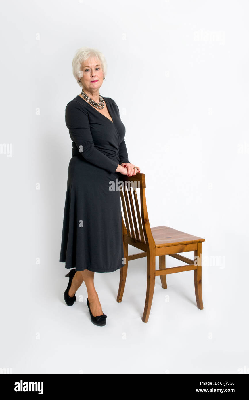 Attractive senior lady stood leaning against wooden chair taken against a white background Stock Photo