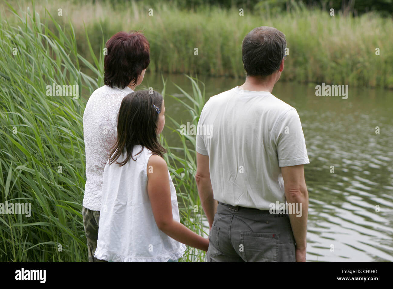 Back view of parents and one only child holding hands and contemplative at water side. Stock Photo