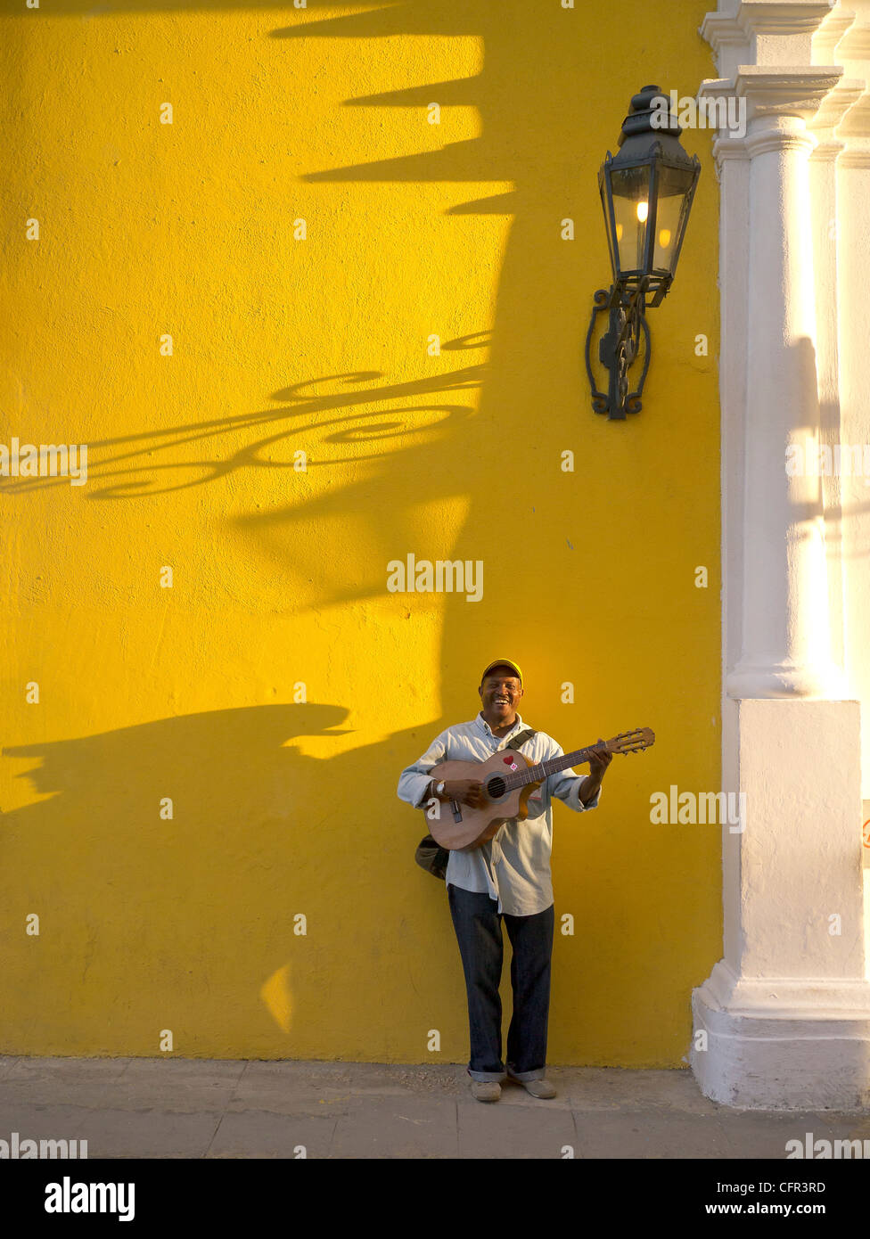 A male Hispanic street musician plays his guitar against a yellow wall late afternoon with strong shadows in Havana, Cuba. Stock Photo