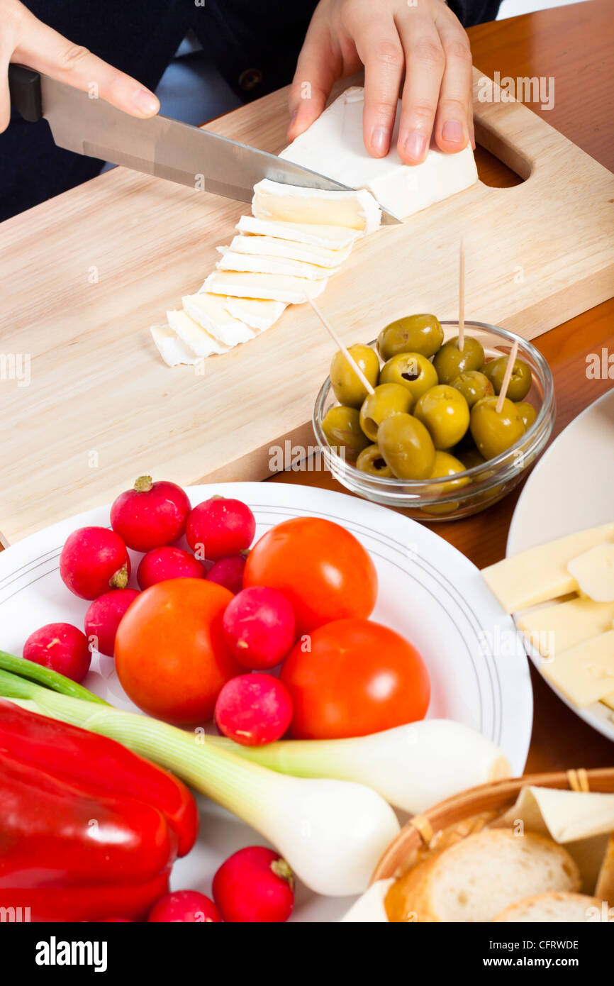 Detail of kitchen table with fresh vegetable, olives and female hands cutting camembert cheese. Stock Photo