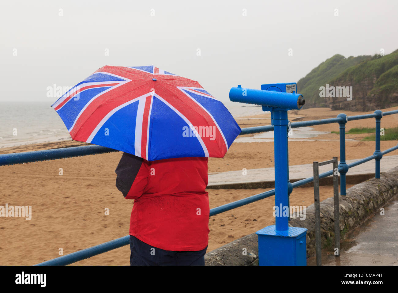 A woman looks out to sea on sea front of deserted seaside with a Union Jack umbrella during very wet British summer weather. Wales UK Great Britain Stock Photo
