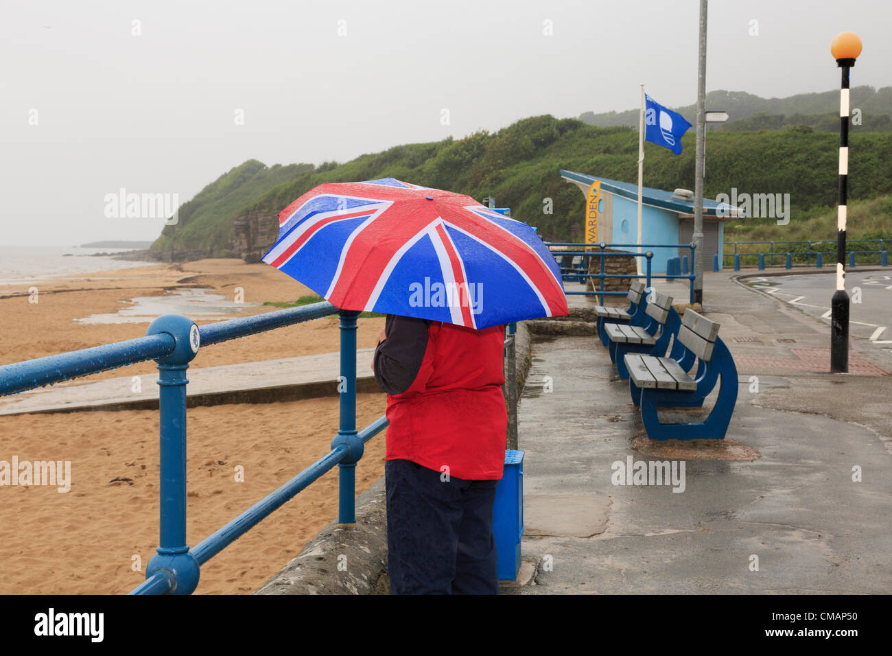 A woman looks out to sea on sea front of deserted seaside with a Union Jack umbrella during very wet British summer weather. UK Stock Photo