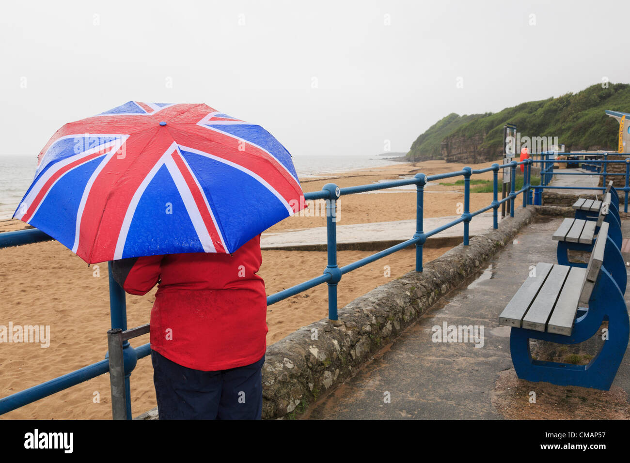 A woman looks out to sea on seafront of deserted seaside with a Union Jack umbrella during very wet British summer weather. UK Britain Stock Photo