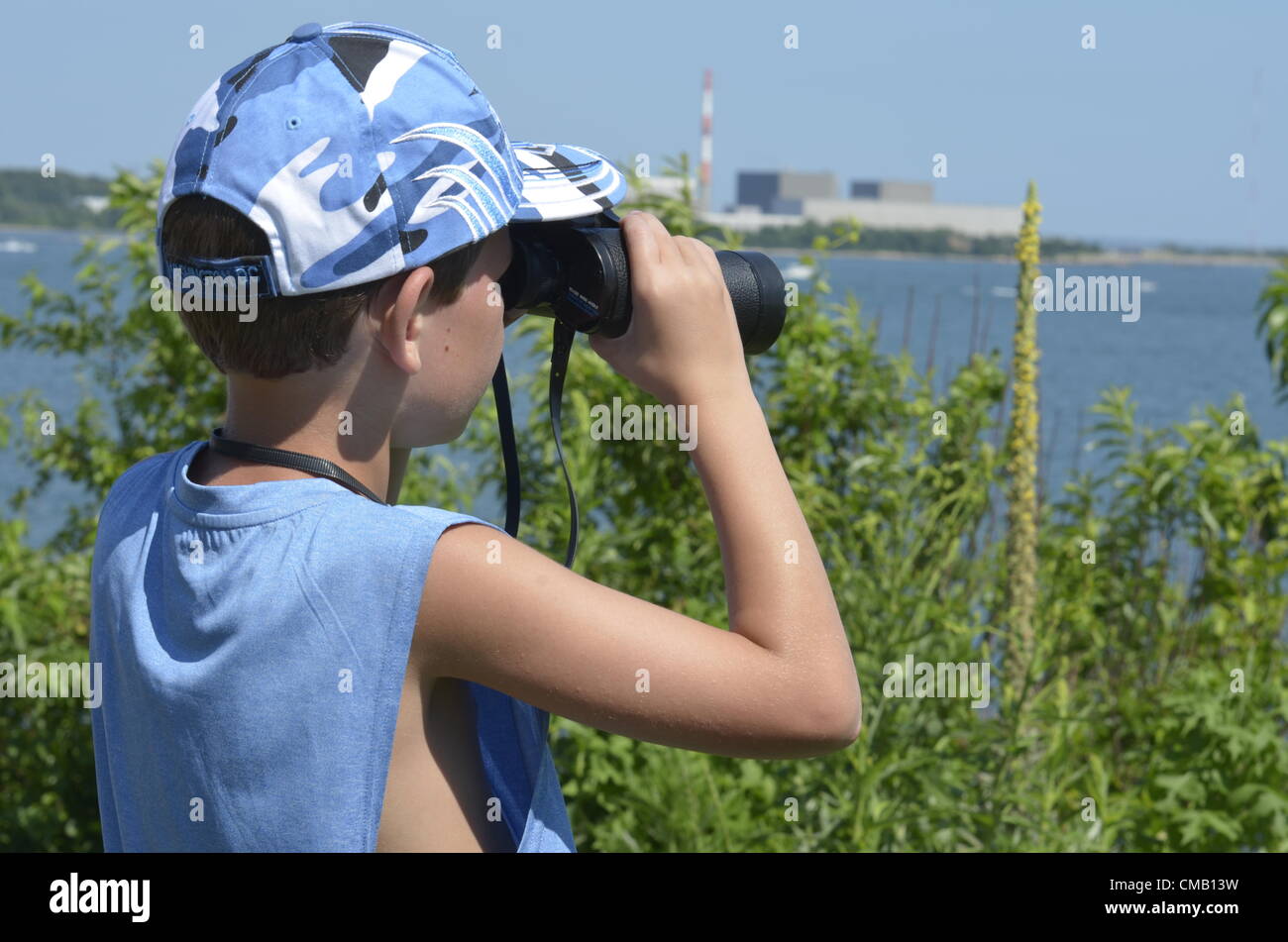 Young explorer. Niantic, Connecticut, USA, July 6, 2012 - From a high vantage point at McCooks Point Park, seven-year-old Nathanial "Nate" Tirrell of Southbury, Connecticut wearing a blue military fatigue style cap looks out to sea with binoculars as some of the tall ships begin to appear in Niantic Bay in East Lyme during OpSail 2012 CT to spot tall ships, both military and civilian. The Millstone Nuclear Power Plant is in the distance. Model released. Stock Photo