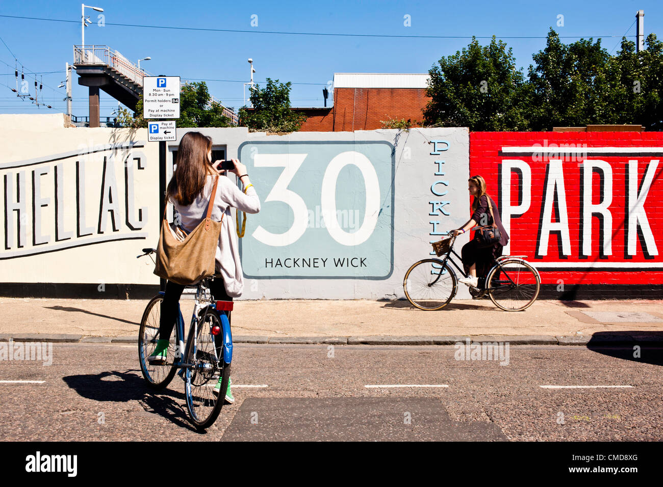 London, UK - 23 July 2012: Two italian tourists take pictures by a graffiti wall in Hackney Wick, close to the London 2012 Olympic Park site. The area, once a major  manufacturing center in the UK, is now a mix of old warehouses converted into artist’s studio and industrial yards attracting now both tourists and Olympic staff members. Stock Photo