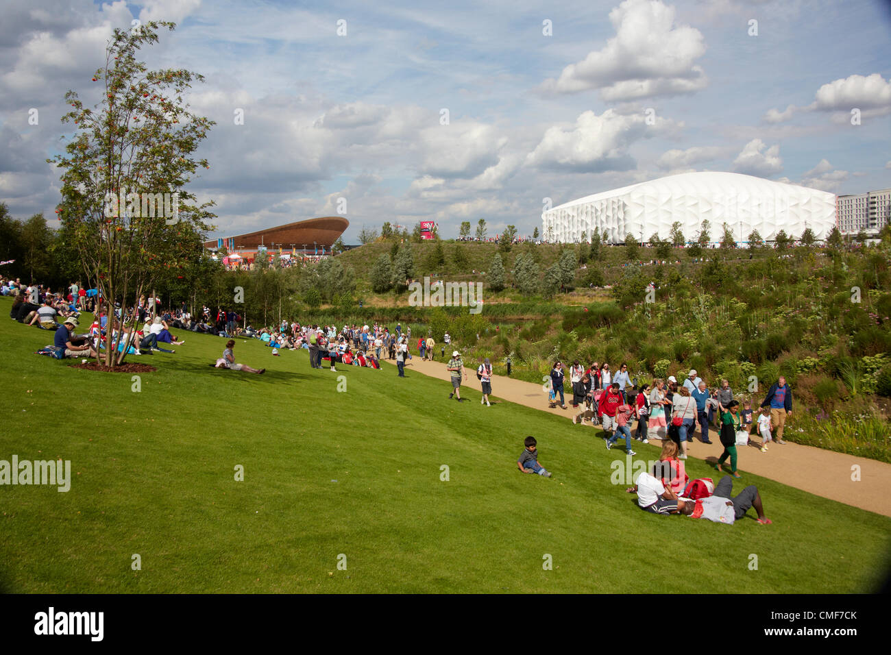 People sitting on lawns in sunshine on a sunny day with Velodrome and Basketball Arena at Olympic Park, London 2012 Olympic Games site, Stratford London E20 UK, Stock Photo