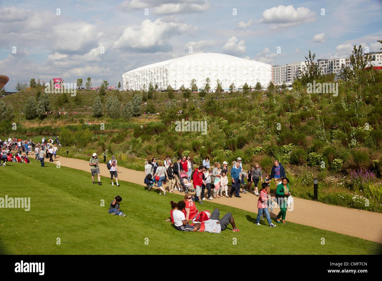 People sitting on lawns in sunshine on a sunny day with Basketball Arena at Olympic Park, London 2012 Olympic Games site, Stratford London E20 UK, Stock Photo