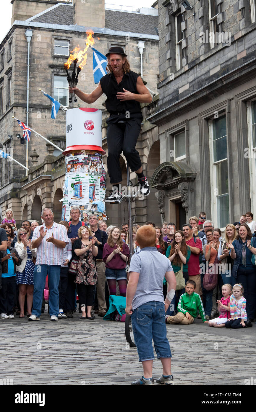 18 August 2012  Damien Ryan, an Australian street performer, riding a unicycle and juggling with fire torches in The Royal Mile, Edinburgh during the Edinburgh Fringe Festival. A small boy who had bee helping him during his show, looks on. Stock Photo