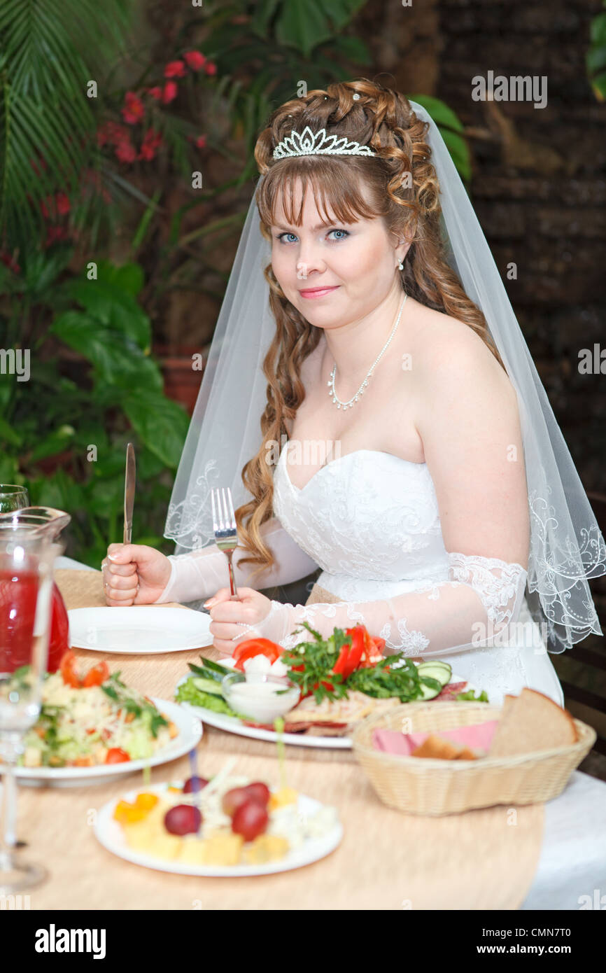 Caucasian bride in white dress sitting at the table with fork and knife ...