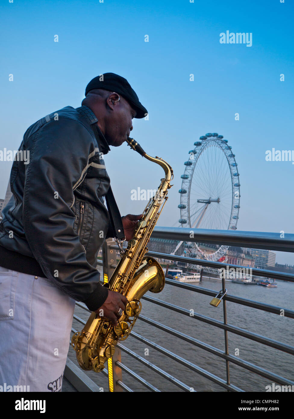Street artist busker playing saxophone on Golden Jubilee Bridge at dusk with River Thames and London Eye in background London UK Stock Photo