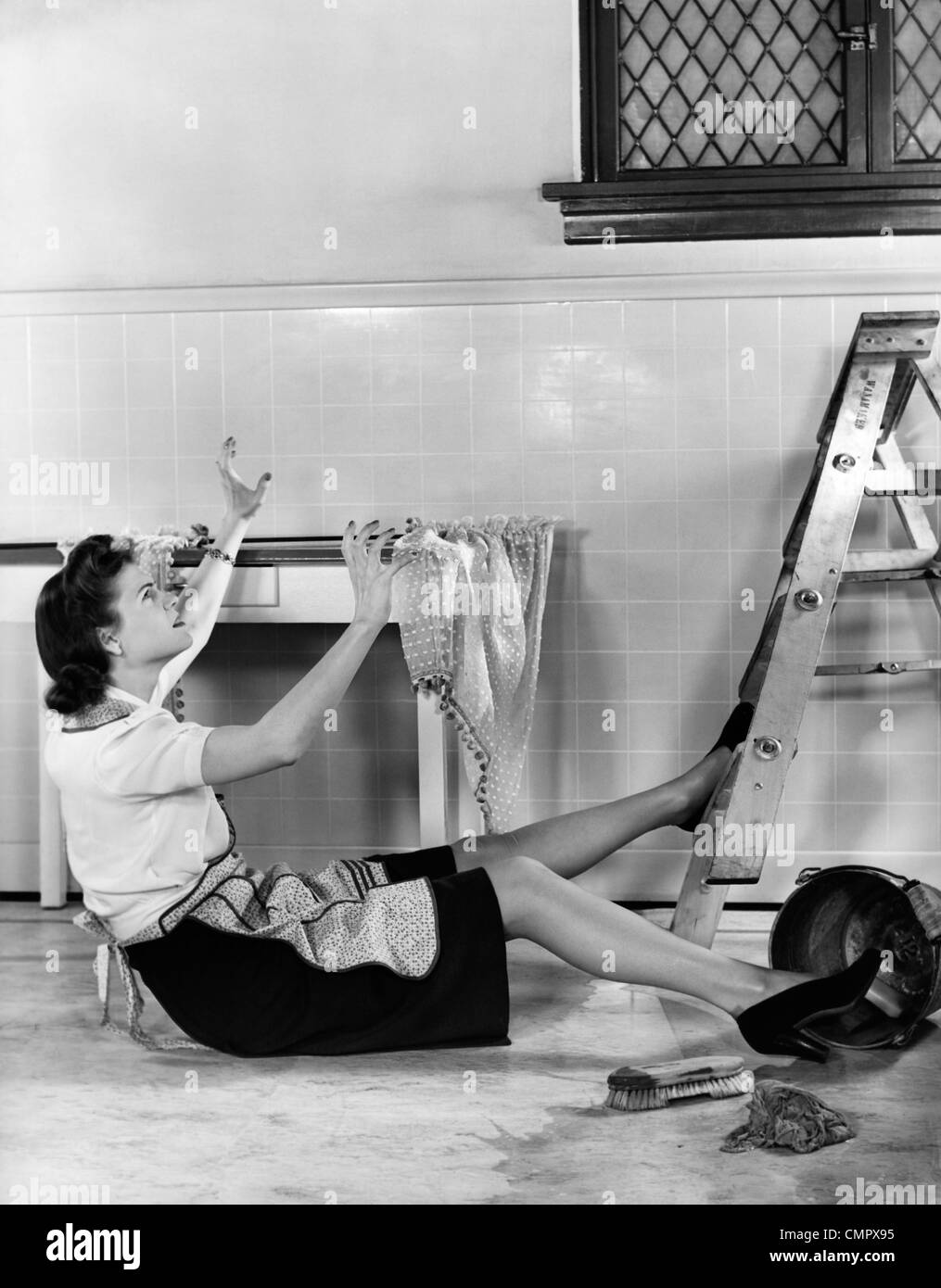 1940s WOMAN HOUSEWIFE FALLING FROM STEP LADDER IN KITCHEN WHILE WASHING WINDOW Stock Photo
