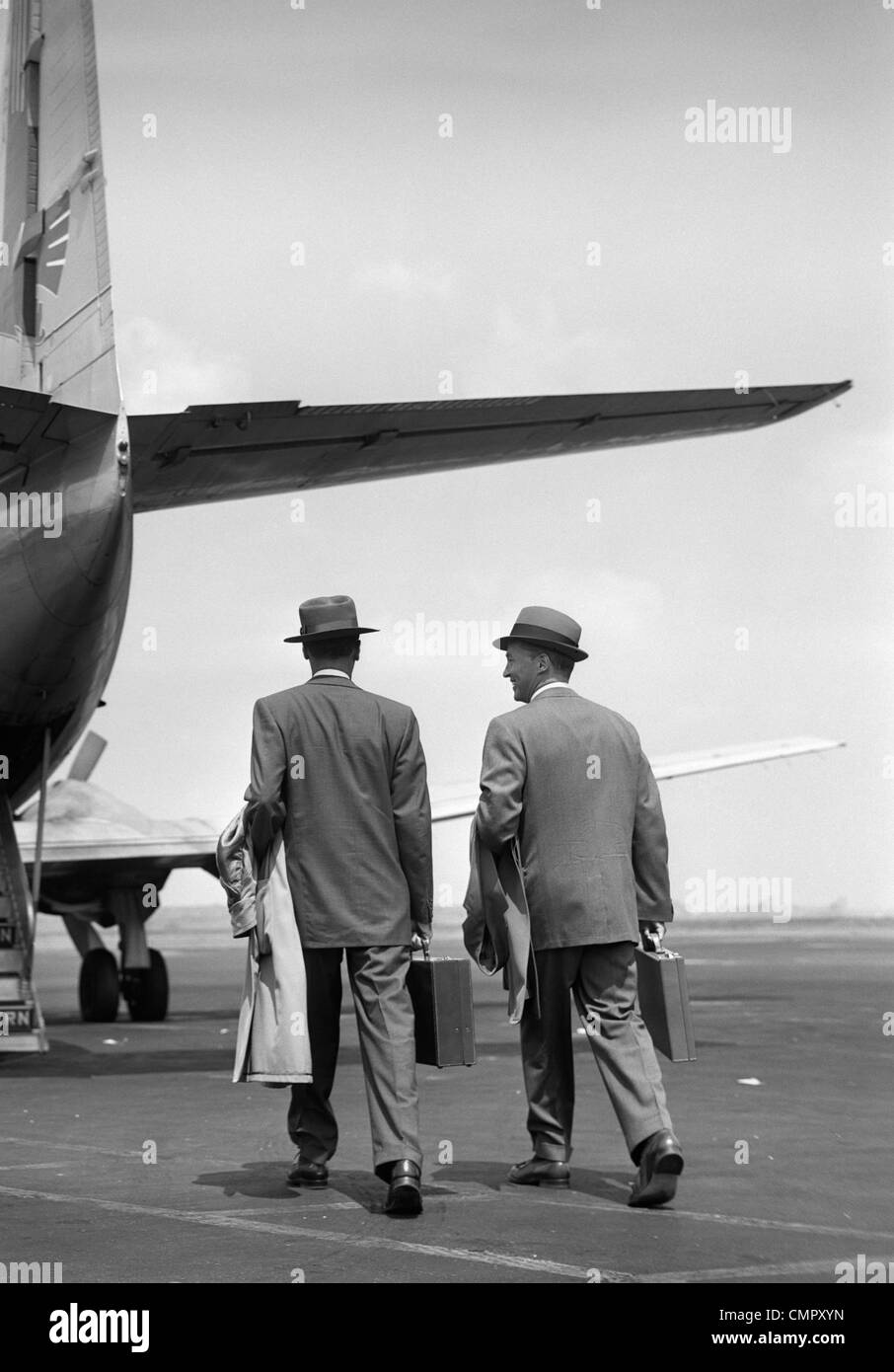 1950s TWO MEN BUSINESSMEN WEARING HATS CARRYING OVERCOATS AND BRIEFCASES WALKING TOGETHER TO BOARD PROPELLER AIRLINER Stock Photo