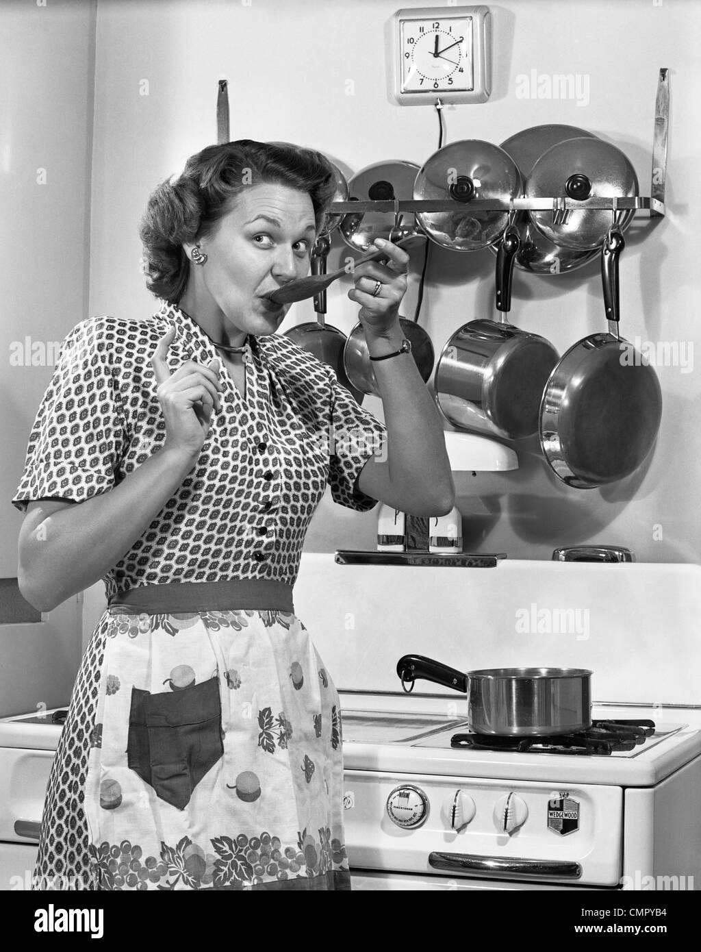 1950s HOUSEWIFE TASTING WITH SPOON BY STOVE FUNNY FACIAL EXPRESSION Stock Photo