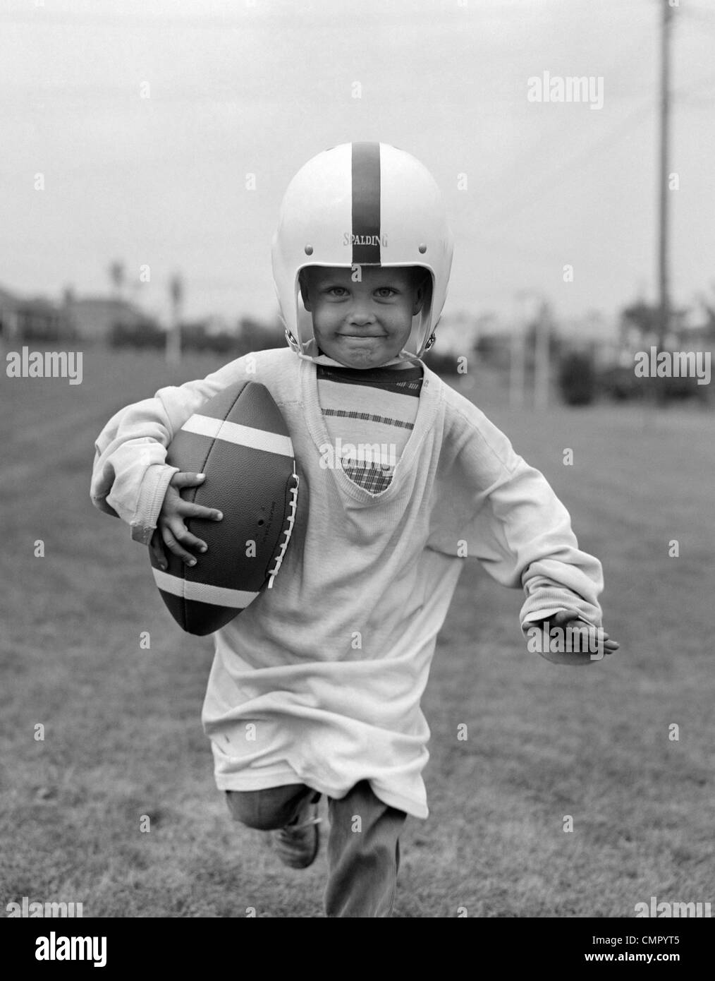 1950s BOY IN OVERSIZED SHIRT & HELMET RUNNING TOWARD CAMERA HOLDING FOOTBALL Stock Photo