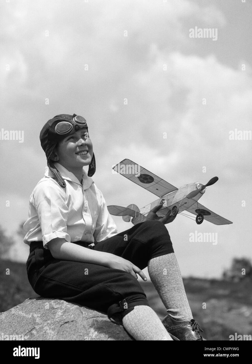 1930s BOY IN LEATHER AVIATOR CAP WITH GOGGLES SITTING ON ROCK HOLDING MODEL PLANE Stock Photo