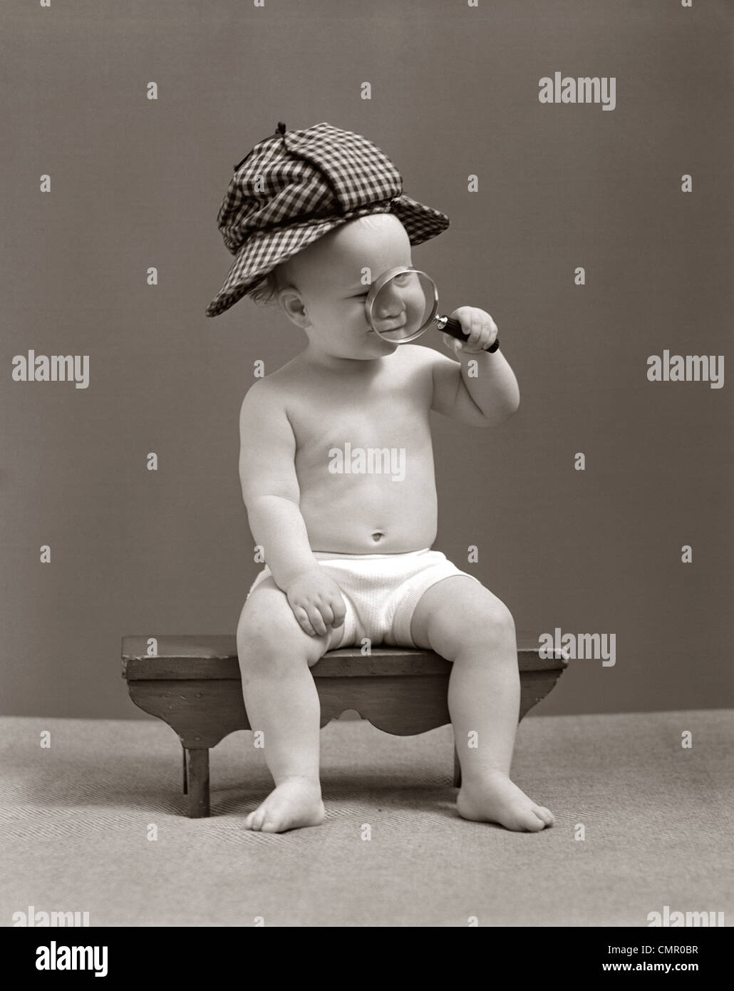 1940s BABY SHERLOCK HOLMES IN DIAPER SITTING ON BENCH WEARING DEER STALKER HAT LOOKING THROUGH MAGNIFYING GLASS Stock Photo