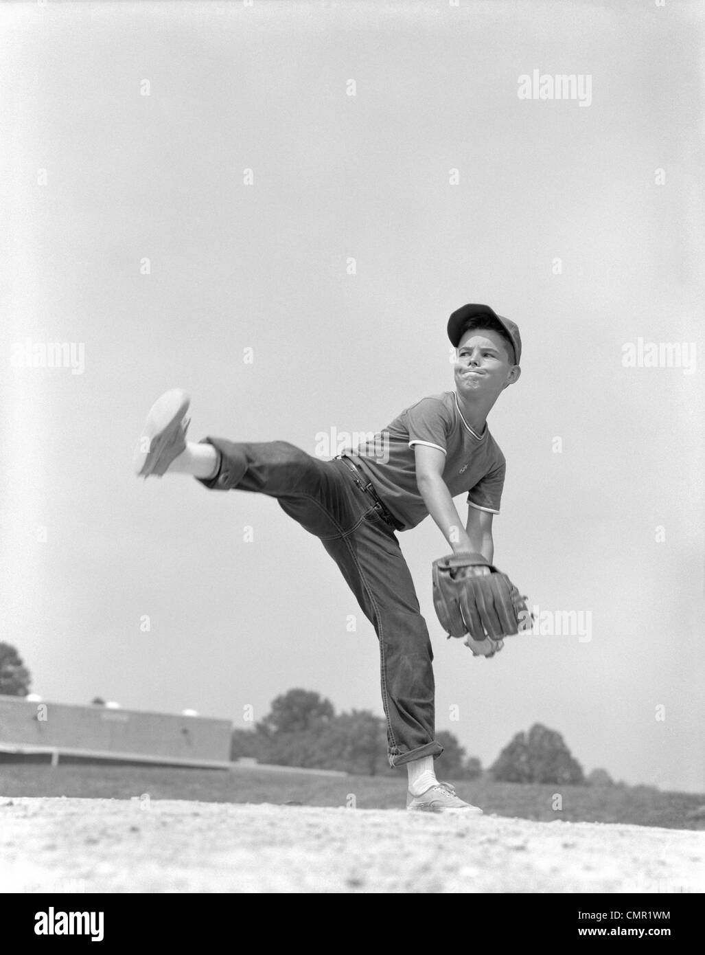 1960s BOY WEARING BASEBALL CAP TEE SHIRT BLUE JEANS AND DETERMINED EXPRESSION IN WIND-UP TO DELIVER PITCH Stock Photo