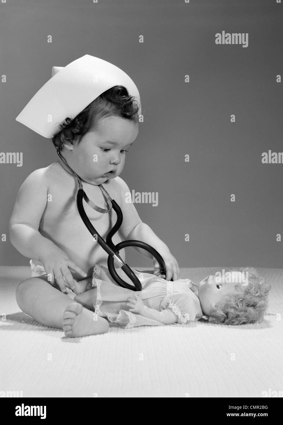 1960s BABY PLAYING NURSE WITH HAT DOLL AND STETHOSCOPE Stock Photo