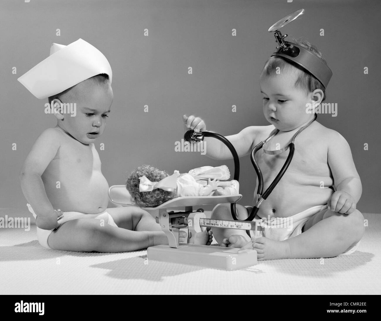 1960s TWO BABIES PLAYING DOCTOR AND NURSE WITH DOLL STUDIO Stock Photo