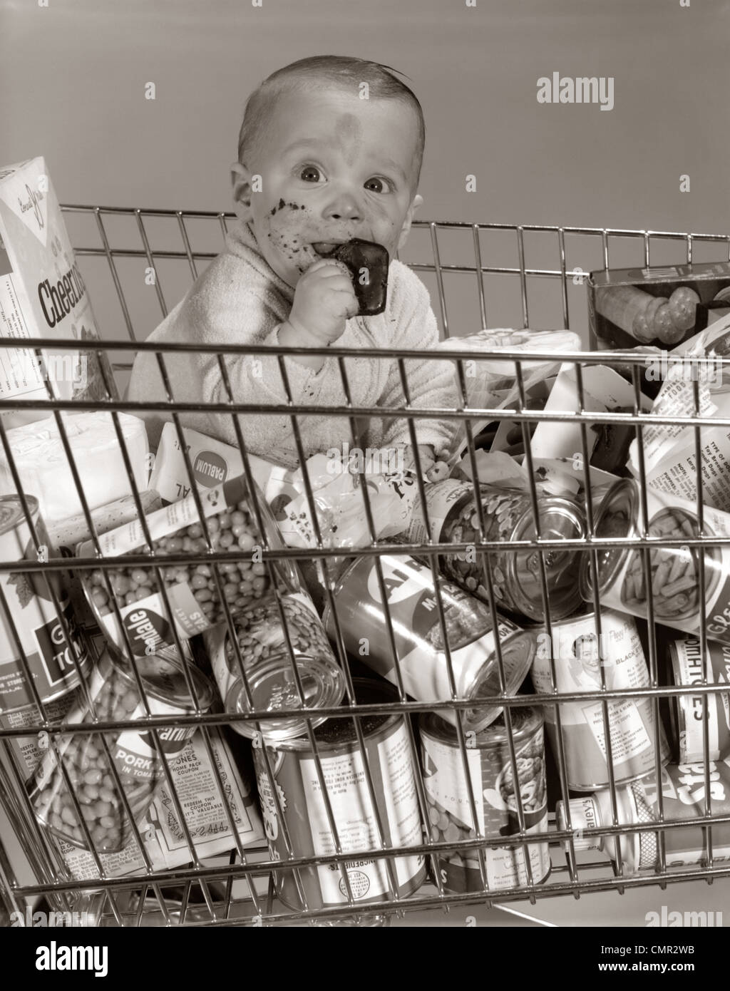 1960s BABY SITTING IN SUPERMARKET CART FULL OF CANS EATING CANDY BAR WITH A MESSY FACE Stock Photo