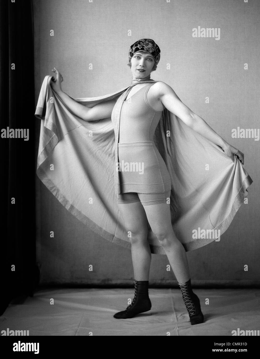 1920s WOMAN IN OLD-FASHIONED BATHING SUIT & CAP HOLDING OUT LARGE CAPE TIED AROUND NECK LOOKING AT CAMERA Stock Photo