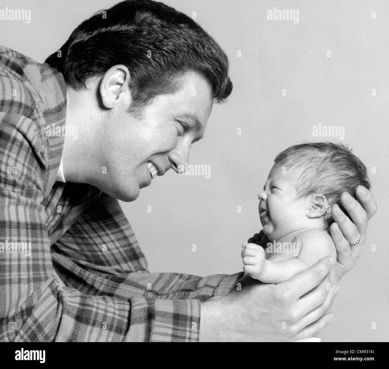 1970s CLOSE-UP PORTRAIT OF SMILING FATHER HOLDING POUTING NEWBORN Stock Photo