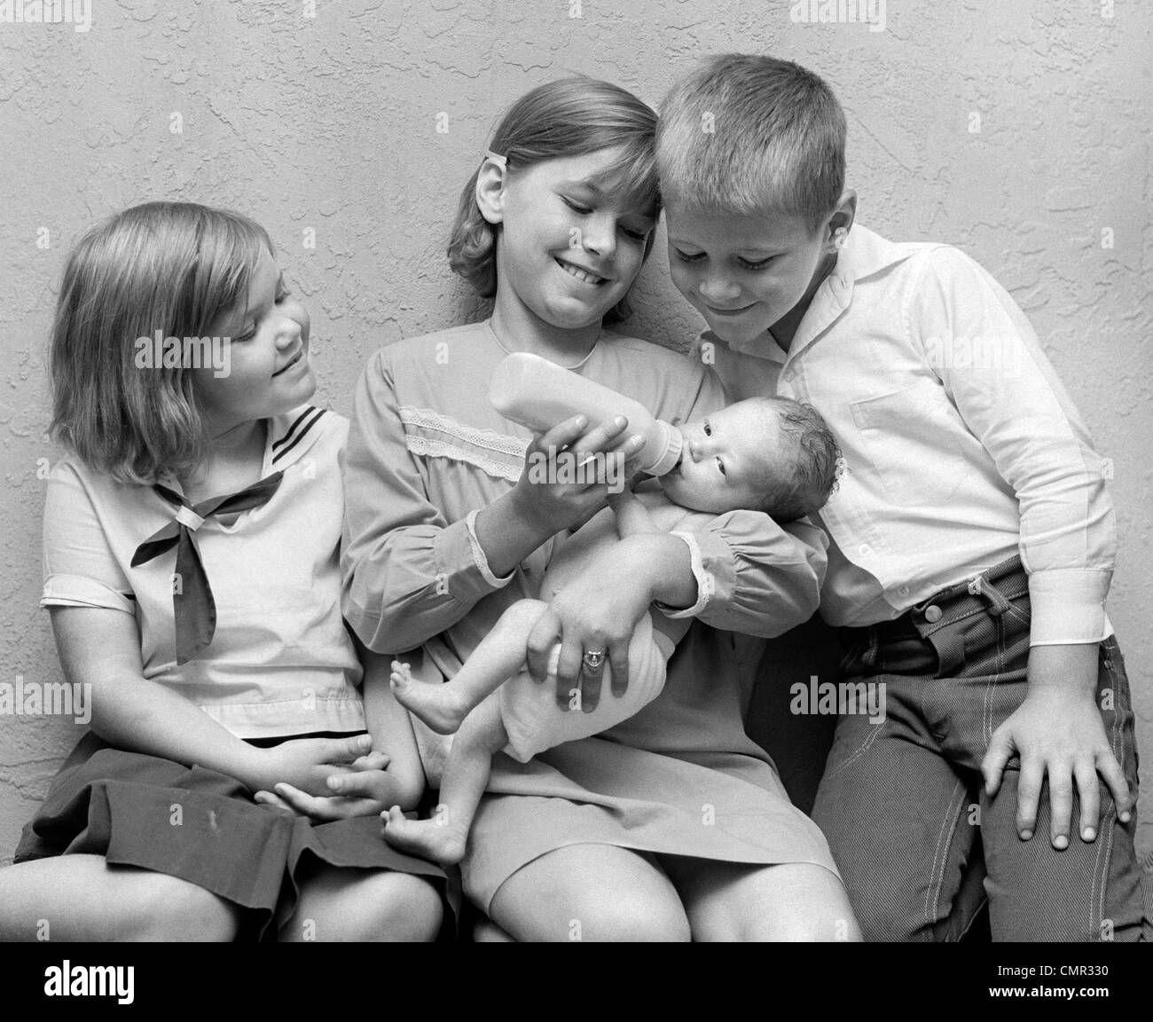 1970s GIRL FEEDING BOTTLE TO BABY SISTER WITH BROTHER & SISTER SITTING