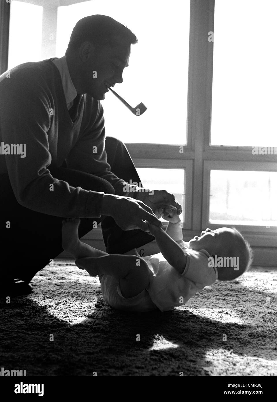 1950s FATHER PIPE IN MOUTH KNEELING DOWN TO PLAY WITH BABY LAYING ON FLOOR SUNLIGHT COMING IN THROUGH WINDOWS Stock Photo