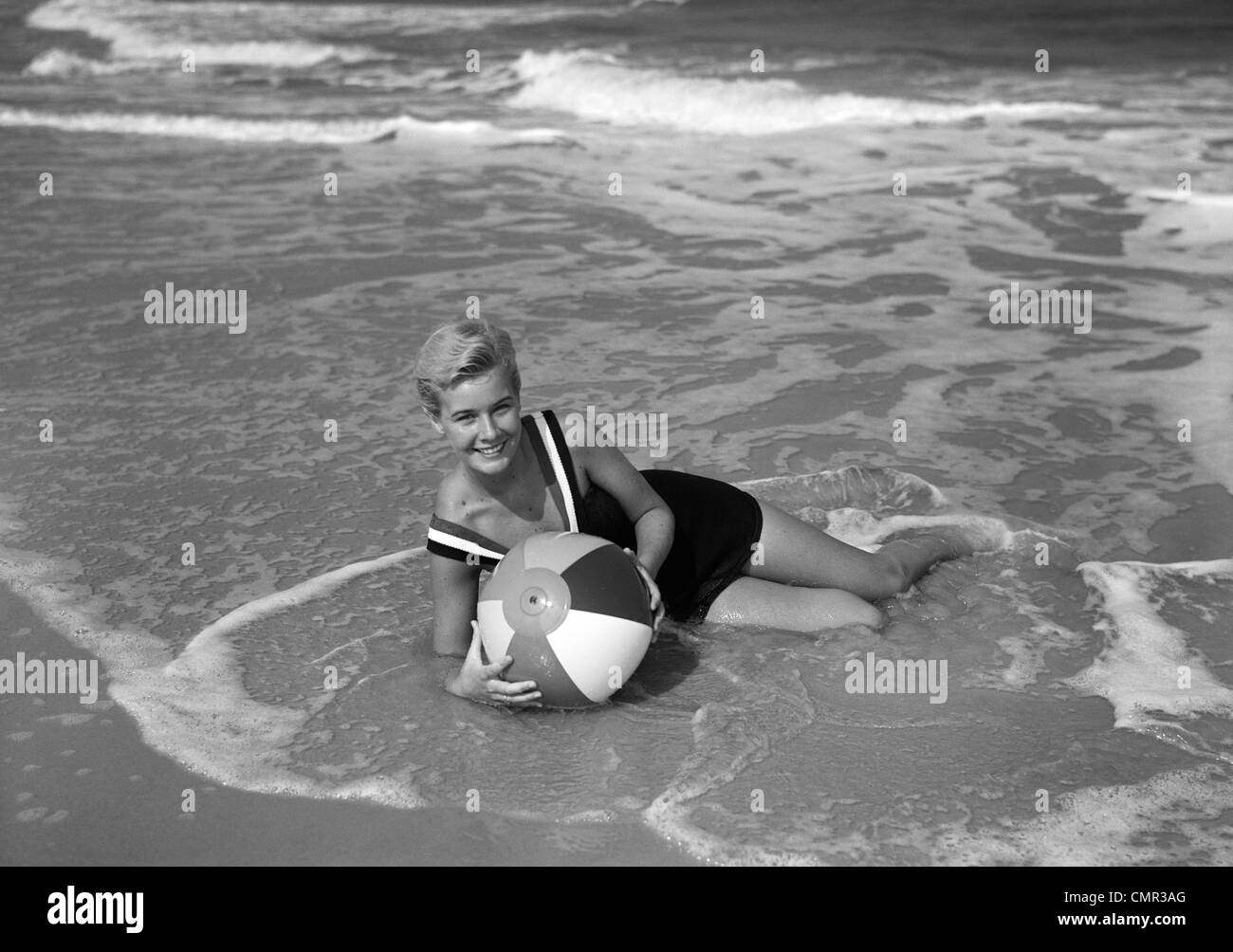 1960s WOMAN IN BATHING SUIT LYING IN THE SURF HOLDING A BEACH BALL SMILING OUTDOOR Stock Photo