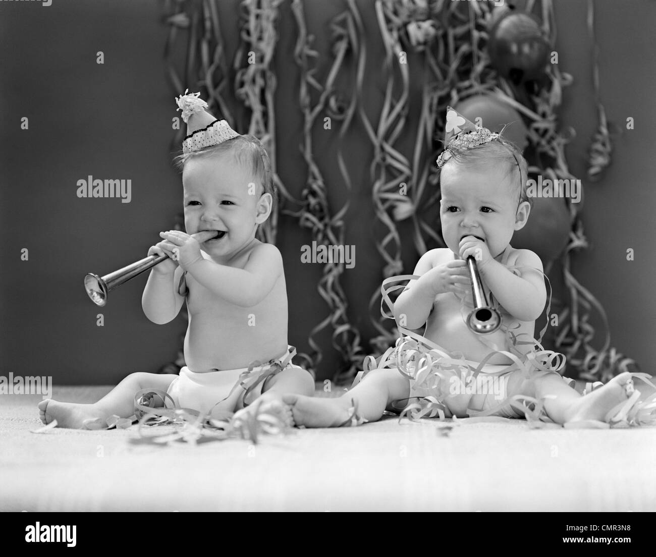 1940s TWIN BABIES WITH PARTY HATS HORNS AND PAPER STREAMERS NEW YEAR CELEBRATION STUDIO Stock Photo