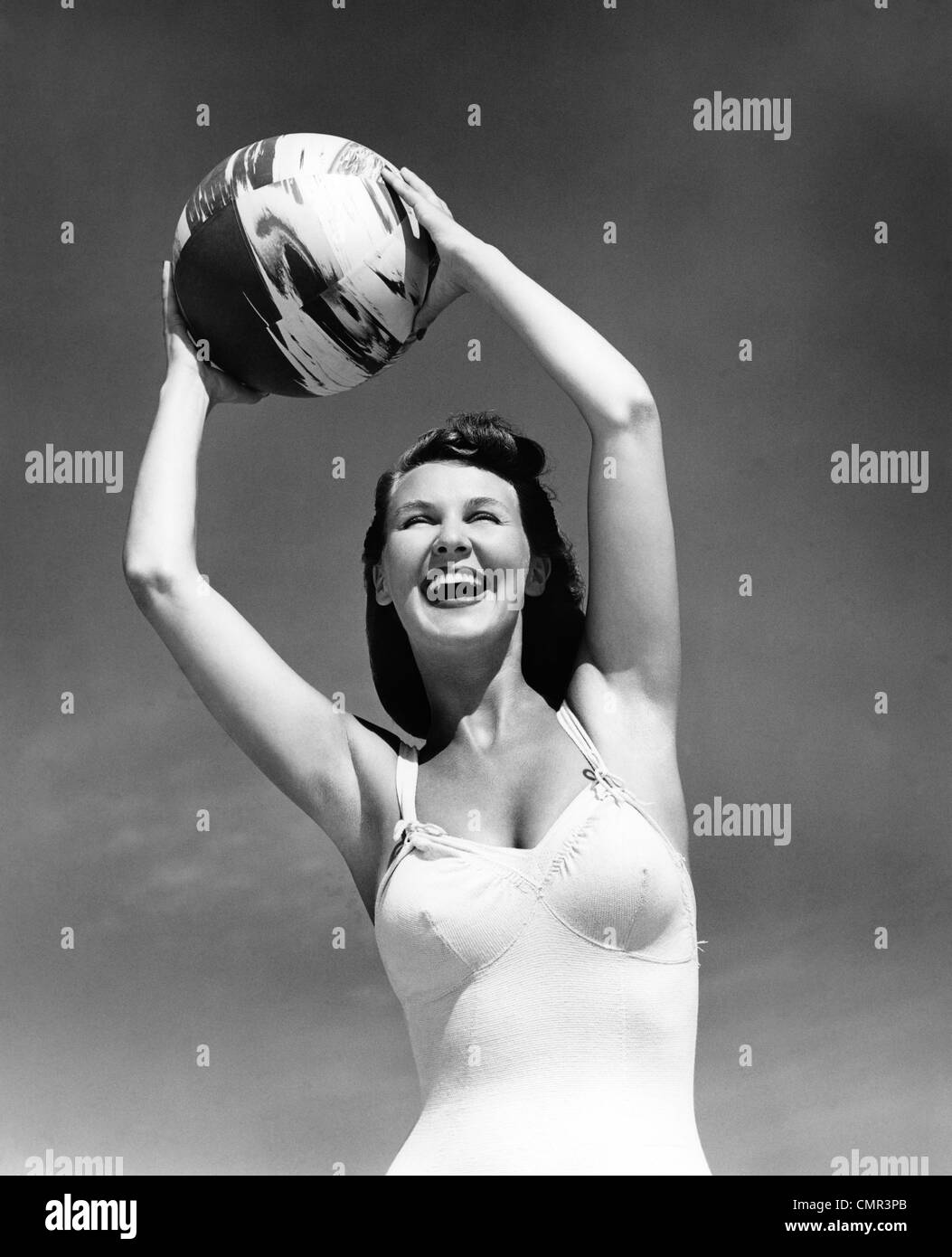 1940s SMILING WOMAN IN WHITE BATHING SUIT HOLDING A BEACH BALL OVER HER HEAD OUTDOOR Stock Photo