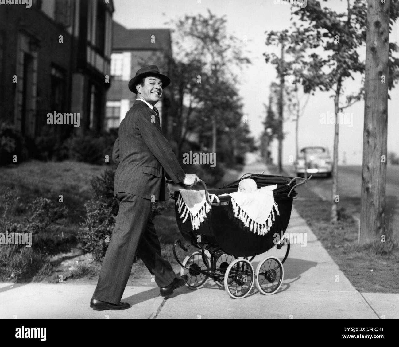 1940s 1950s SMILING FATHER IN SUIT & HAT PUSHING BABY IN CARRIAGE DOWN SIDEWALK LOOKING AT CAMERA Stock Photo