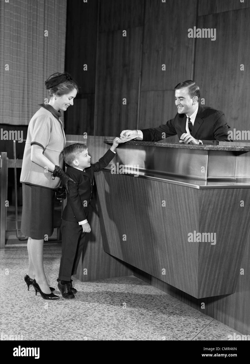 1960s MOTHER IN BANK WITH SON HANDING PASSBOOK TO TELLER Stock Photo