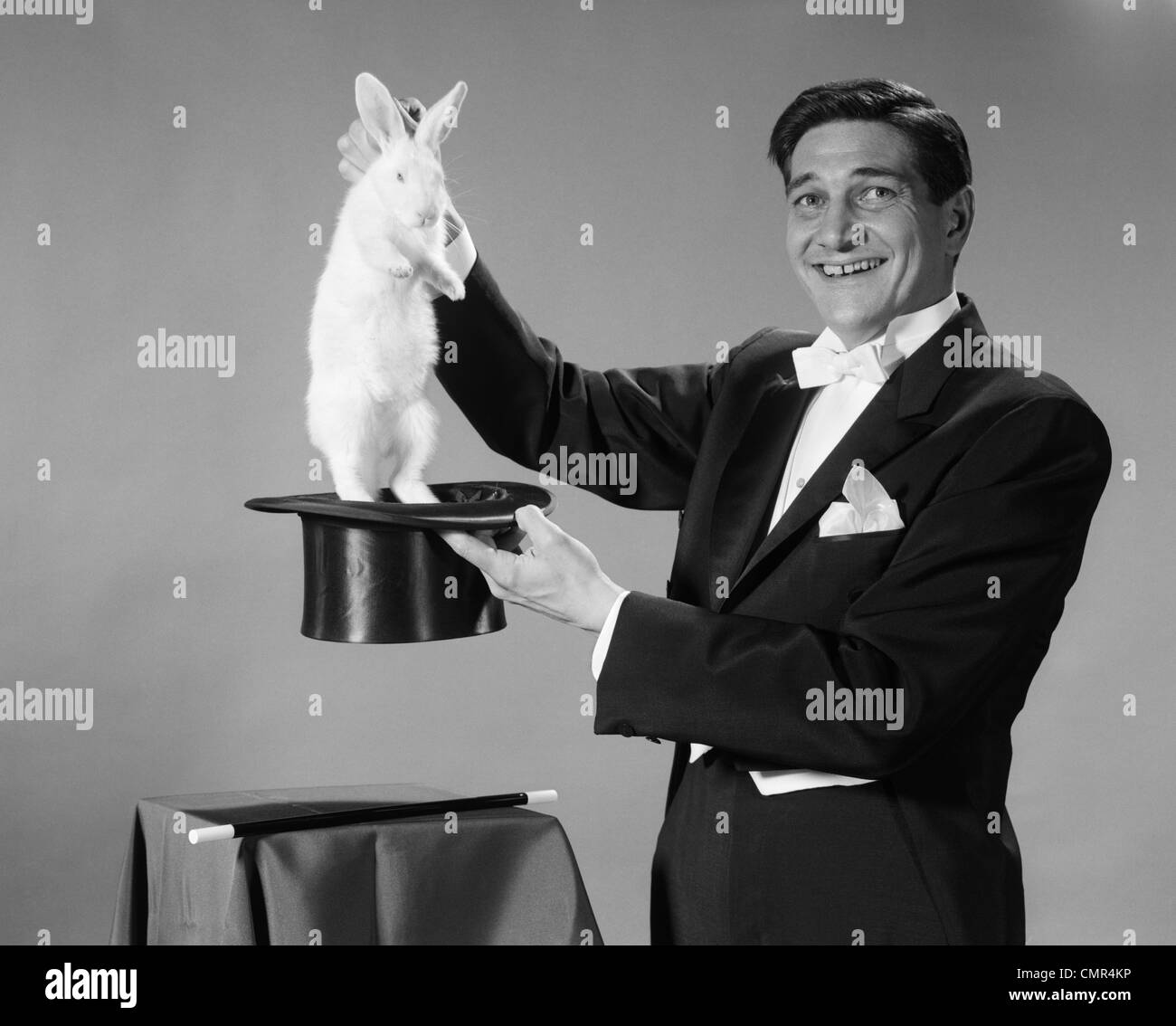 1960s MAN PORTRAIT SMILING MAGICIAN PULLING WHITE RABBIT OUT OF TOP HAT Stock Photo