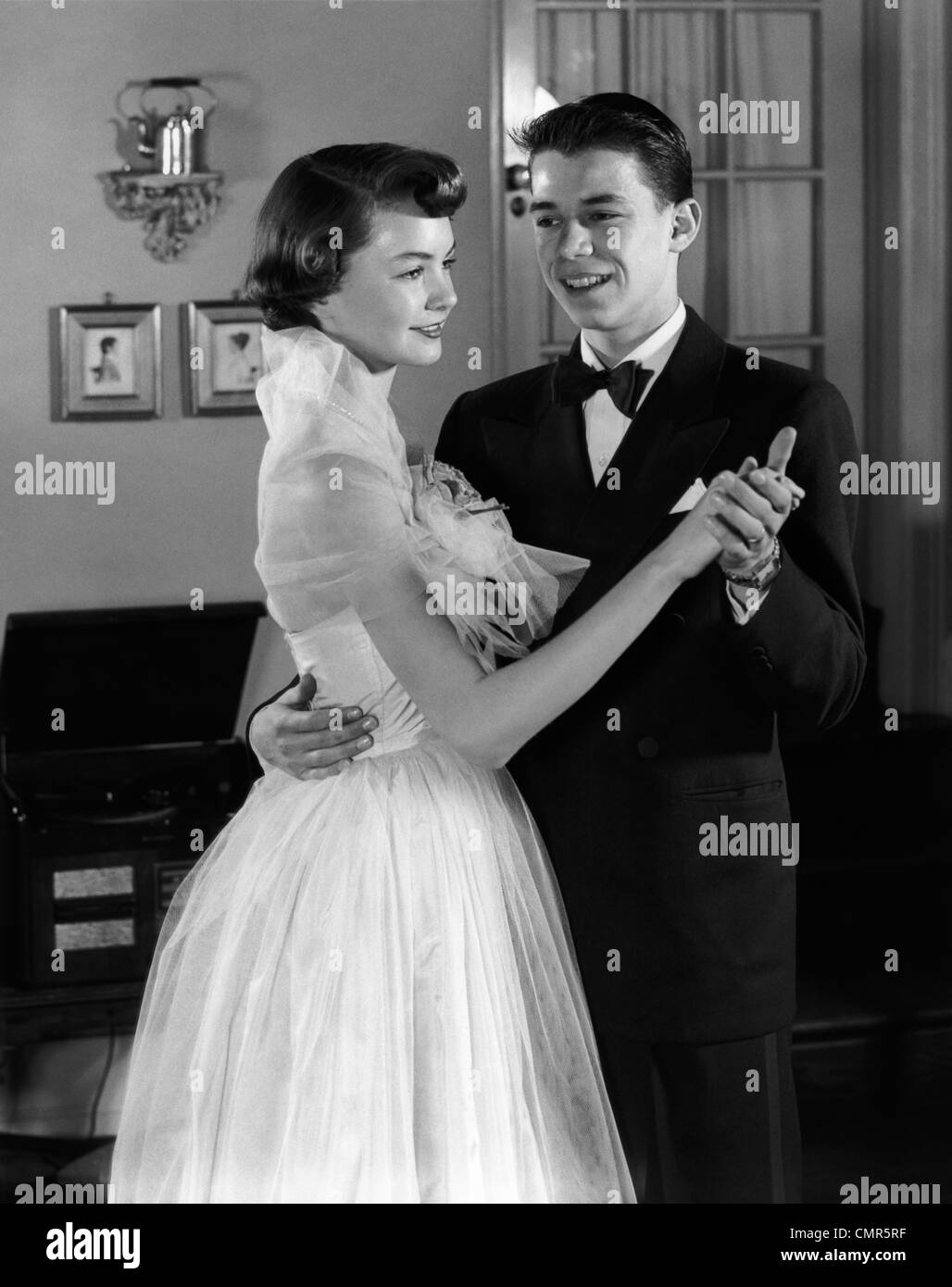 1950s SMILING TEENAGE COUPLE IN FORMAL EVENING WEAR DANCING INDOORS Stock Photo