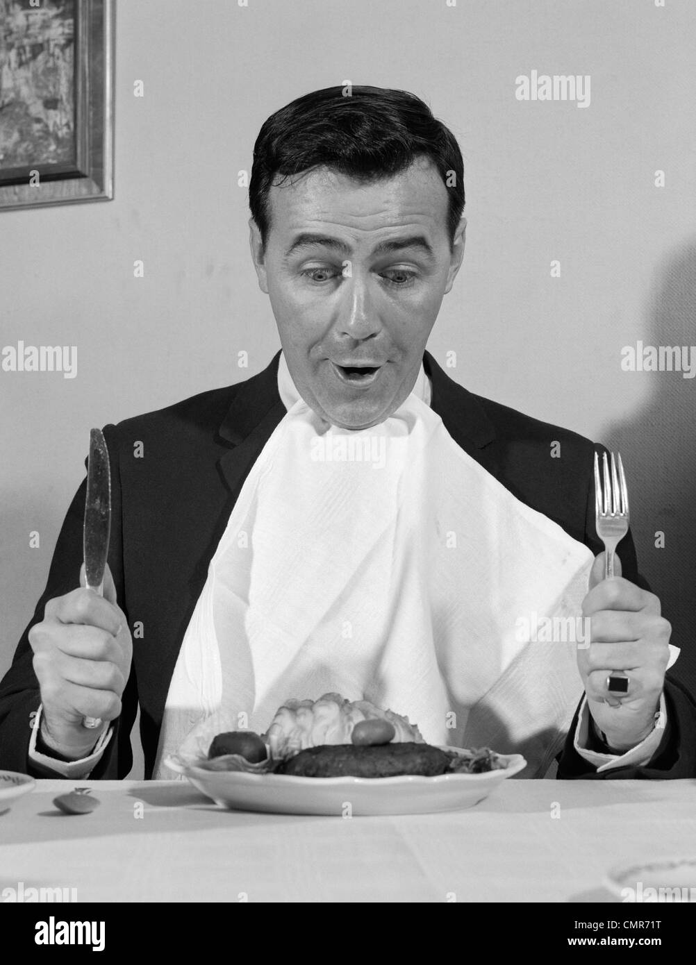 1960s MAN SITTING AT TABLE READY TO EAT DINNER WITH NAPKIN AT NECK AND KNIFE & FORK IN AIR Stock Photo