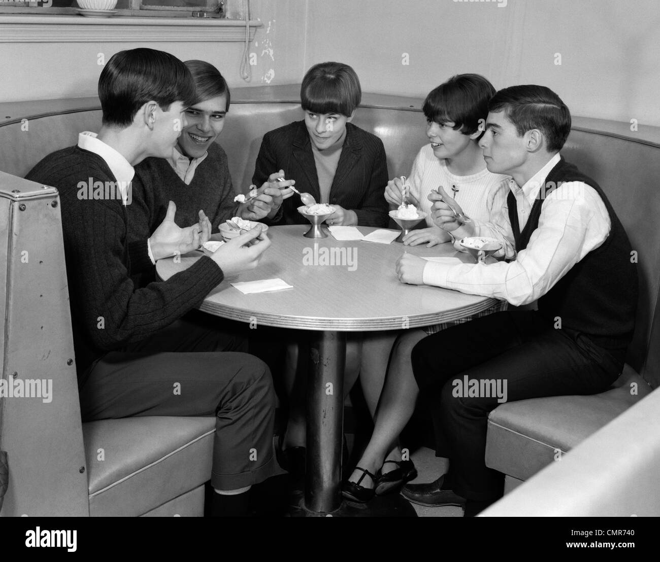 1960s GROUP OF 5 TEENS SITTING AT ROUND BOOTH EATING ICE CREAM SUNDAES & TALKING Stock Photo