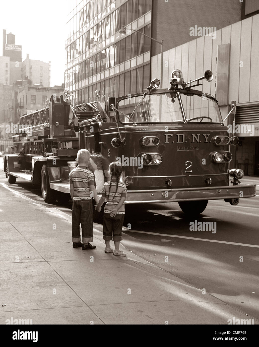 1970s 2 CHILDREN BOY GIRL HOLDING HANDS LOOKING AT FIRE TRUCK PARKED ON STREET NEW YORK CITY Stock Photo