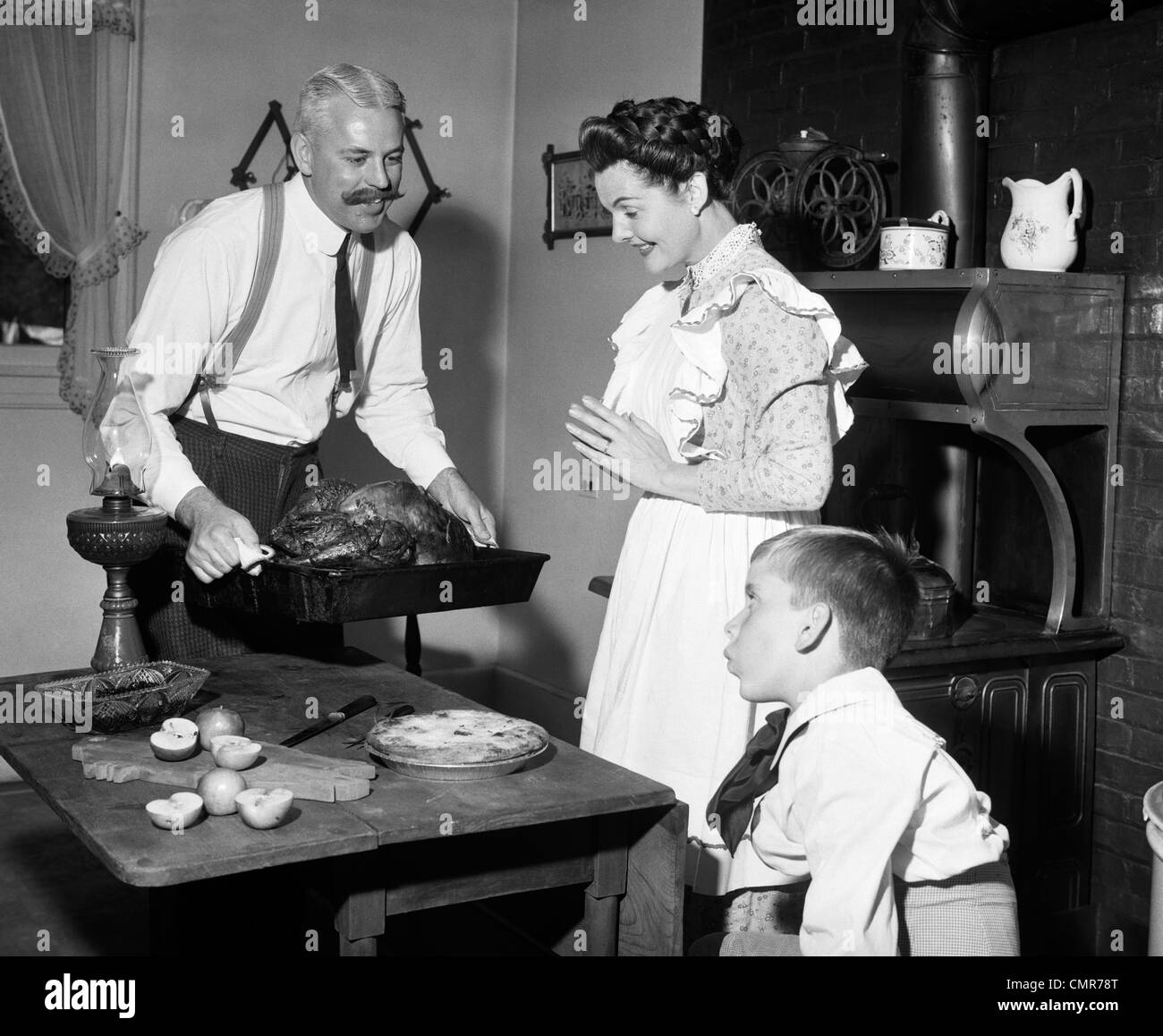 1890s 1900s MOCK TURN OF THE 20TH CENTURY THANKSGIVING DINNER FATHER TAKING TURKEY OUT OF STOVE SHOWING IT TO WIFE AND SON Stock Photo