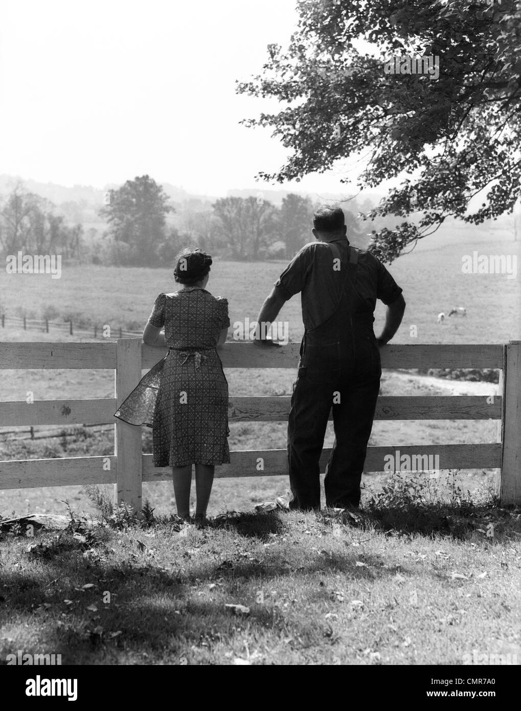 1940s REAR VIEW OF ELDERLY FARM COUPLE STANDING AT FENCE LOOKING OUT ONTO PASTURE Stock Photo