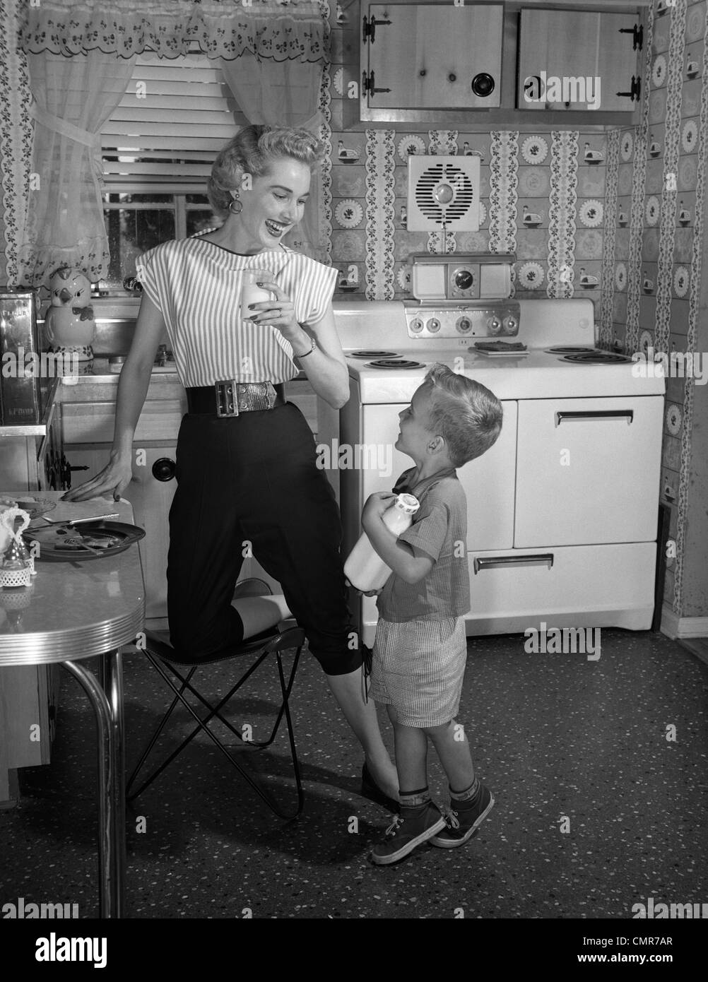 1950s BOY IN KITCHEN HOLDING BOTTLE OF MILK LOOKING UP AT MOTHER DRINKING GLASS OF MILK Stock Photo