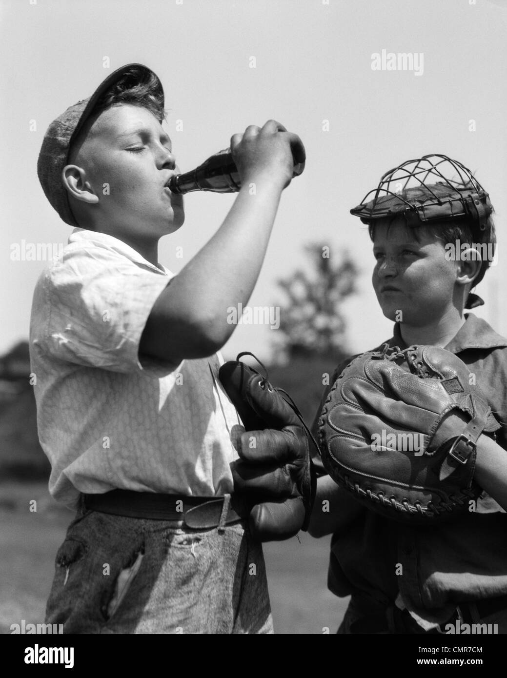 1930s PAIR OF BOYS WEARING BASEBALL GLOVES ONE WITH CATCHER'S MASK WATCHING OTHER WEARING CAP DRINK BOTTLE OF SODA Stock Photo
