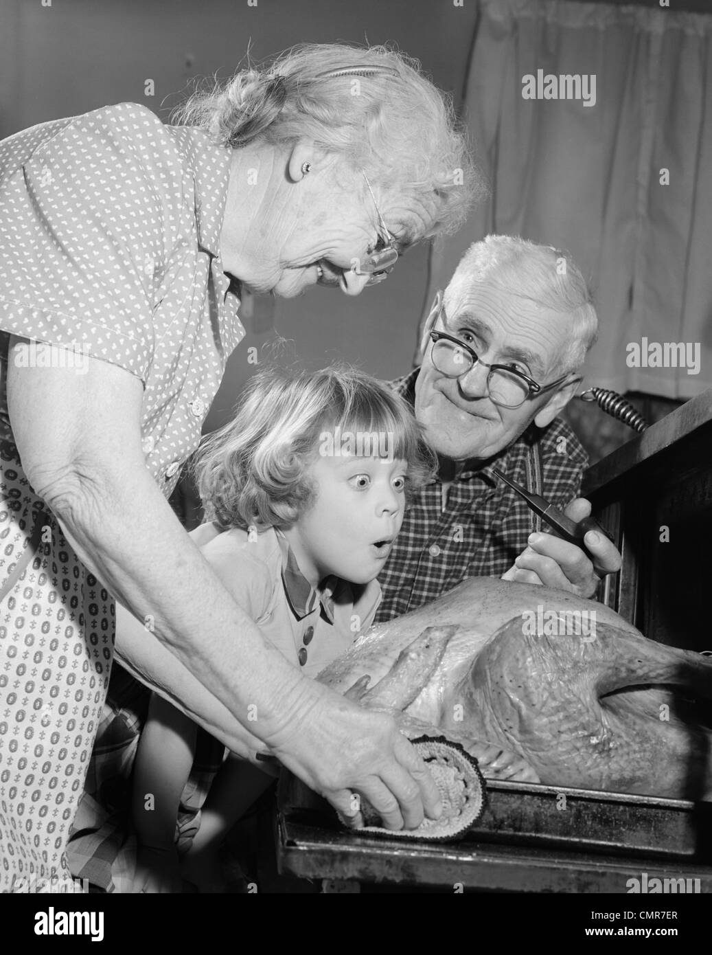 1950s 1960s EXCITED LITTLE GIRL GRANDDAUGHTER WITH GRANDPARENTS WATCHING ROAST TURKEY COMING OUT OF OVEN Stock Photo