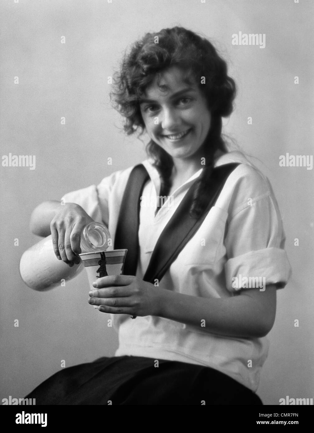 1920s SMILING YOUNG WOMAN SITTING POURING MILK FROM BOTTLE INTO CUP LOOKING AT CAMERA Stock Photo
