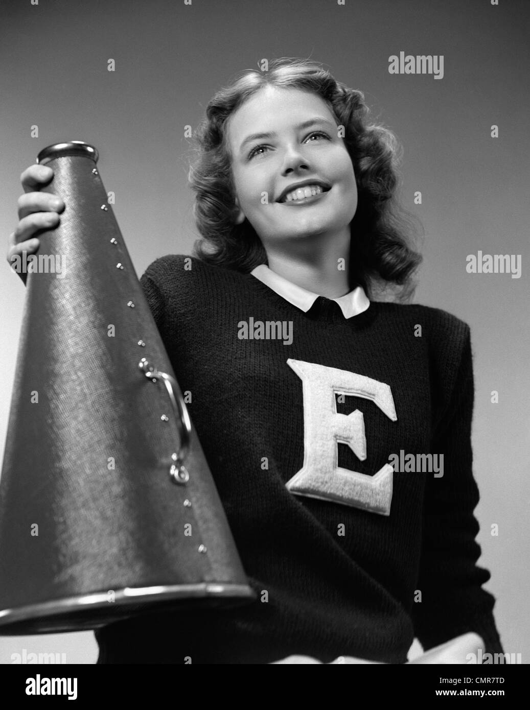 1940s SMILING GIRL WEARING A VARSITY SWEATER WITH THE LETTER E ON FRONT HOLDING A MEGAPHONE WITH HER RIGHT HAND Stock Photo
