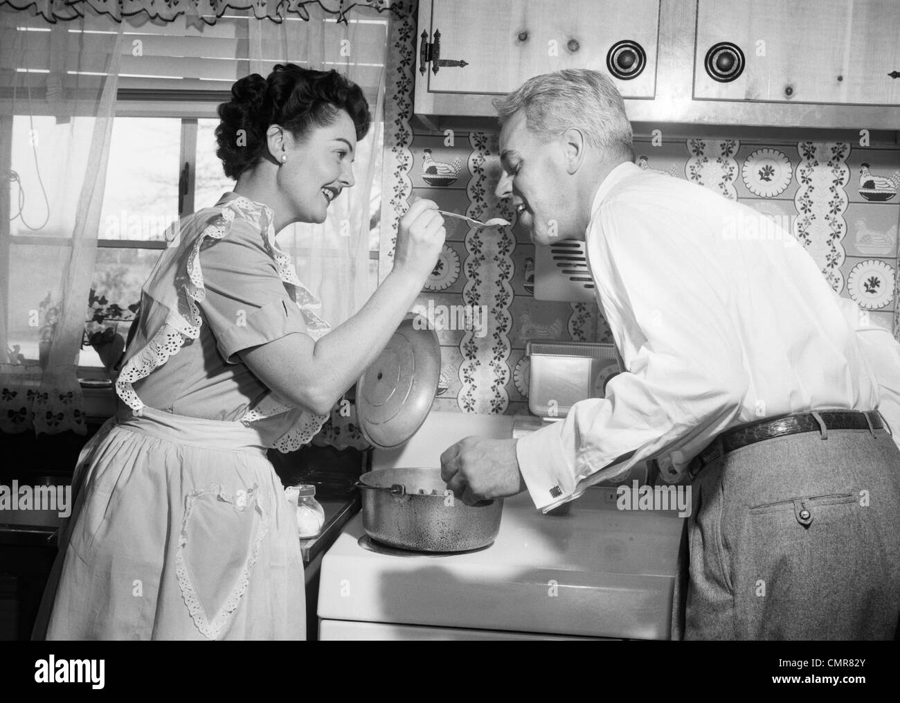 1950s HOUSEWIFE IN KITCHEN HAVING HUSBAND TASTE FOOD ON STOVE Stock Photo
