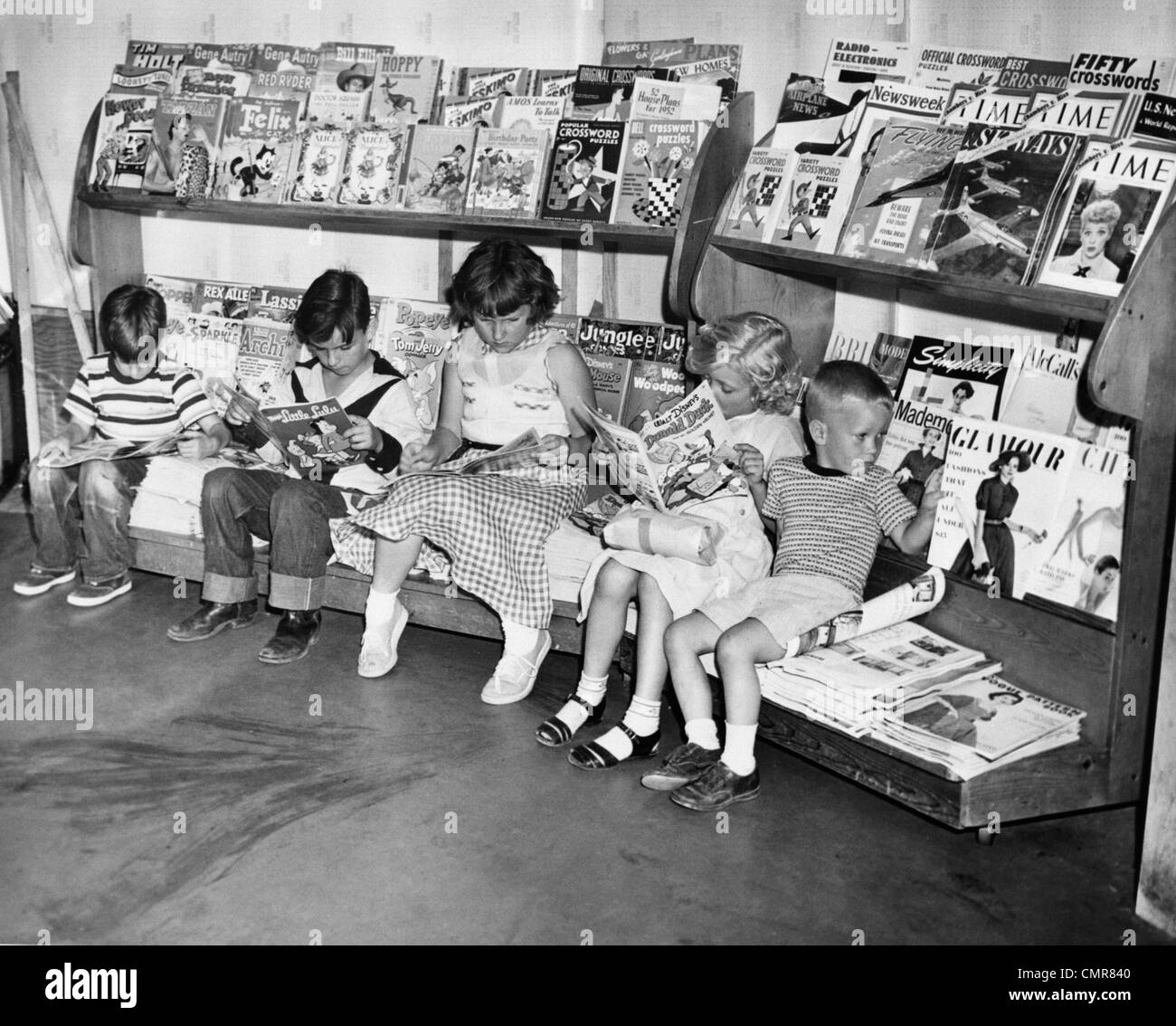 1950s GROUP OF 5 CHILDREN SITTING ON NEWSSTAND READING MAGAZINES COMIC BOOKS Stock Photo