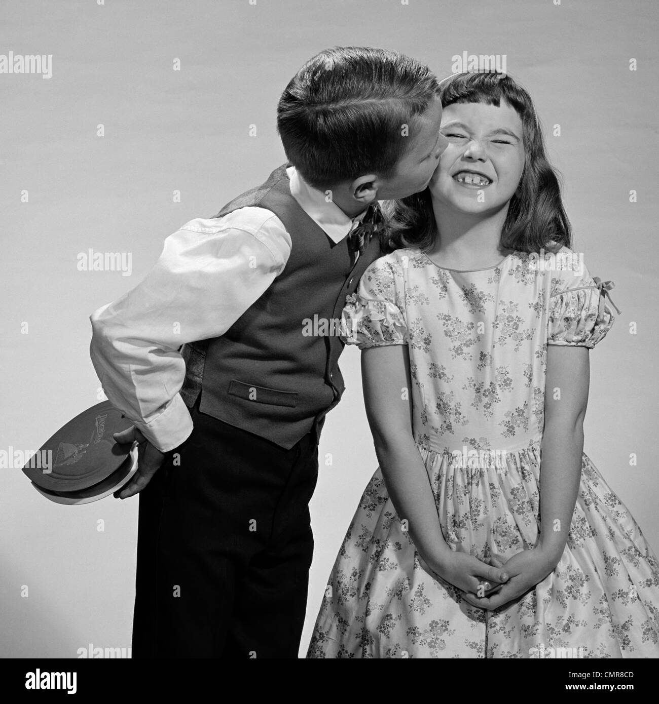 1950s 1960s BOY IN VEST & BOW TIE HOLDING VALENTINE CANDY KISSING CHEEK OF GIRL MAKING A FACE Stock Photo