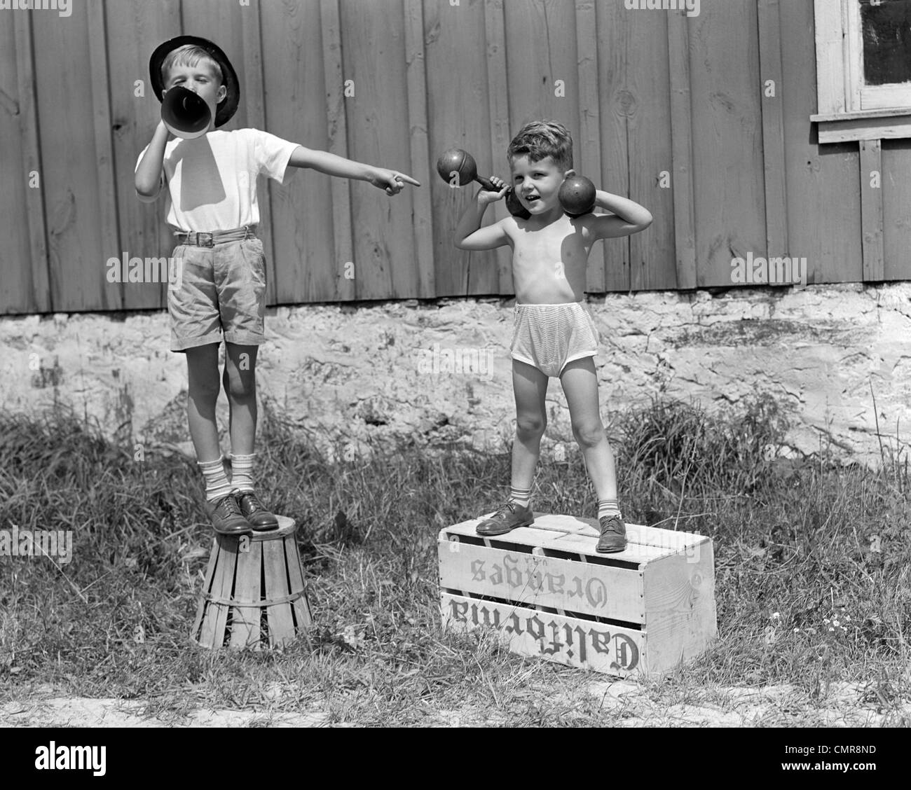 1930s 1940s BOYS PLAYING CARNIVAL STRONGMAN ONE LIFTING DUMBBELLS OTHER ANNOUNCING ACT THROUGH MEGAPHONE Stock Photo