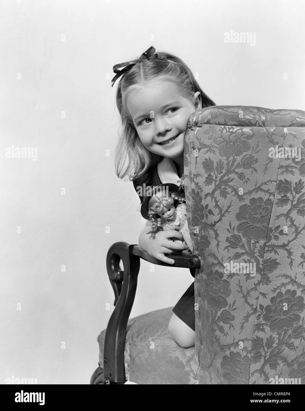 1940s LITTLE GIRL HOLDING DOLL PEEKING AROUND SIDE OF CHAIR Stock Photo